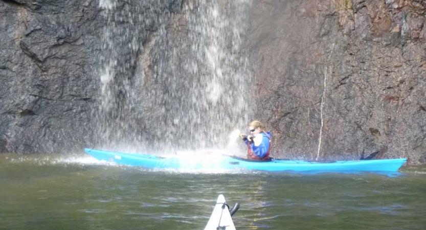 a person in a blue kayak paddles under a waterfall in front of a rock wall